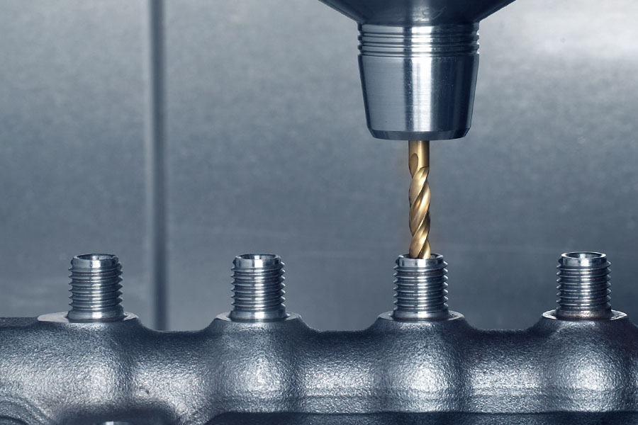 A twist drill is drilling a hole in a cast part that already has several drilled holes and threads.
