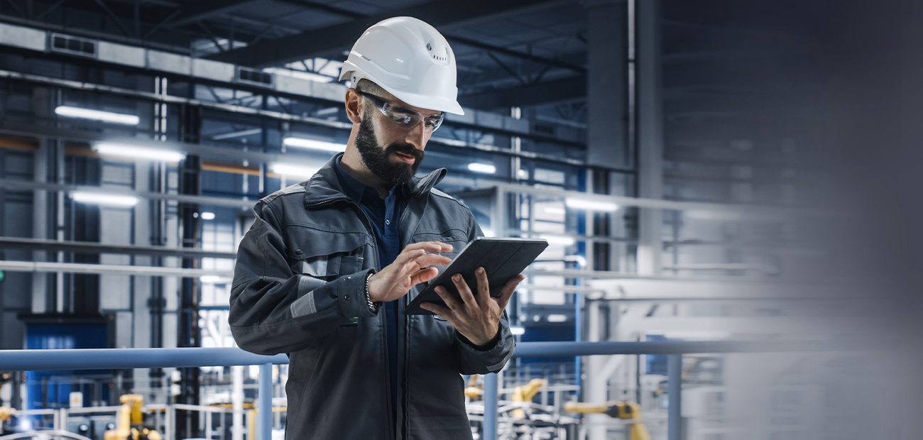 Ein Ingenieur, der in einer modernen Produktionsumgebung mit MRO Verantwortung ein Tablet verwendet. Er trägt einen Schutzhelm und -brille