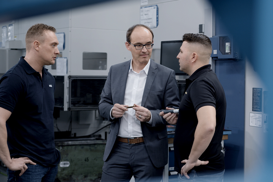 Drei Männer in einem industriellen Arbeitsumfeld, inmitten von Maschinen, die anscheinend eine Diskussion führen