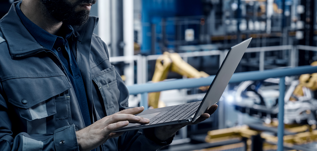 Ingenieur in einer Werkhalle verwendet ein Laptop zur Navigation in einer digitalen Lernwelt, umgeben von automatisierten Maschinen