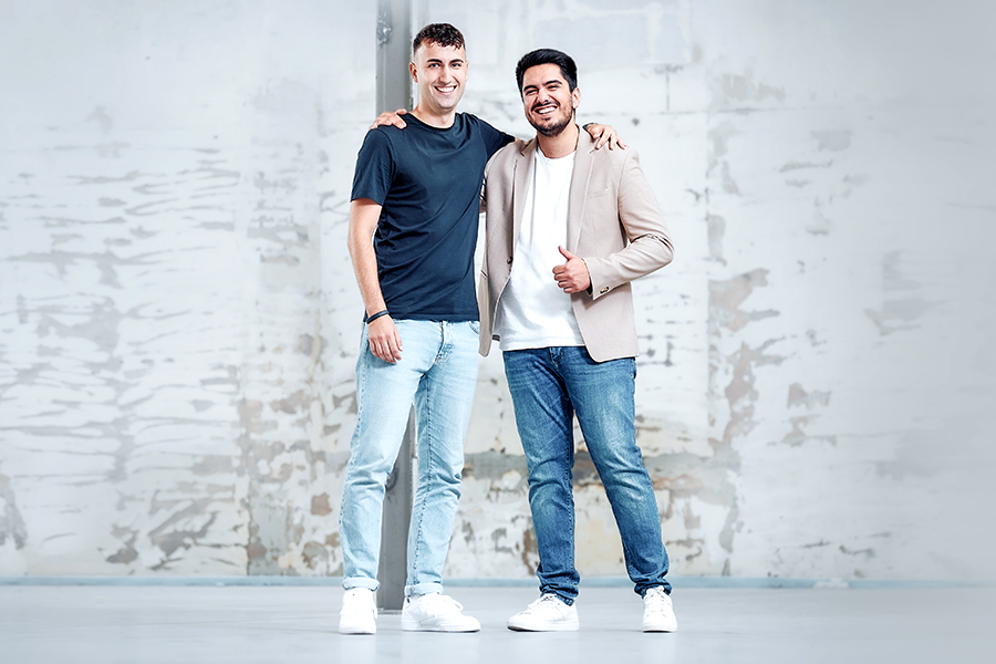 Two young men stand in front of a rustic wall and smile at the camera.