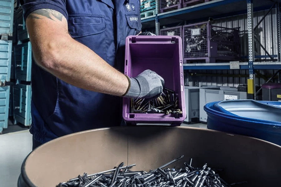 Workers in blue overalls and gloves fill used metal drill bits into a purple recycling box in an industrial workshop