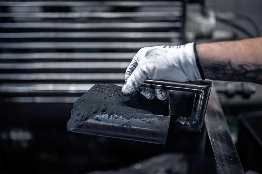 A worker in protective gloves holds a shovel full of tungsten carbide powder in an industrial environment