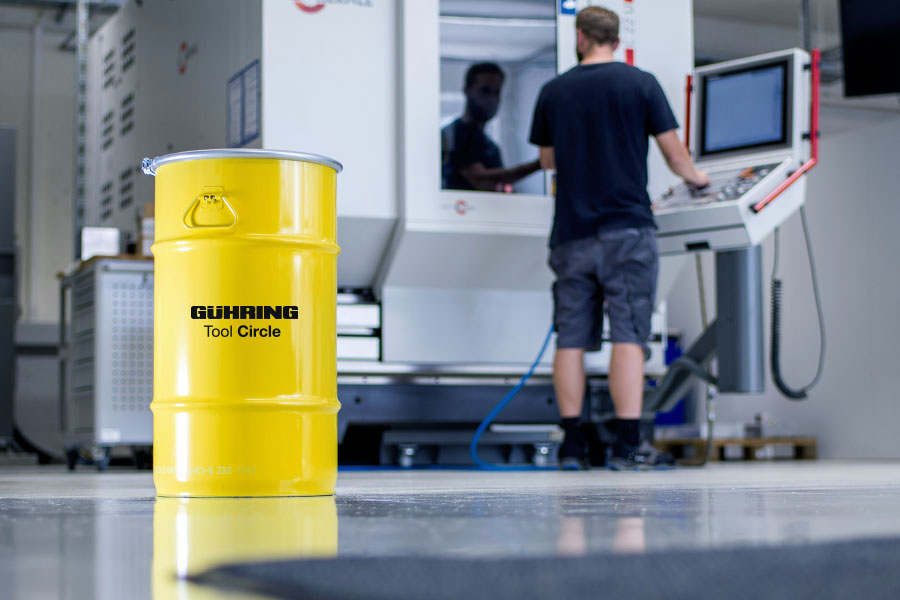 Yellow recycling bin in the foreground of an industrial room, while a technician works on a modern CNC machine in the background