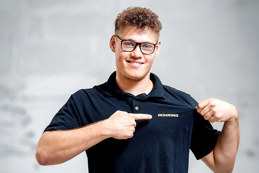 Young man in work clothes with glasses, smiling and proudly pointing at his employer's logo on his polo shirt