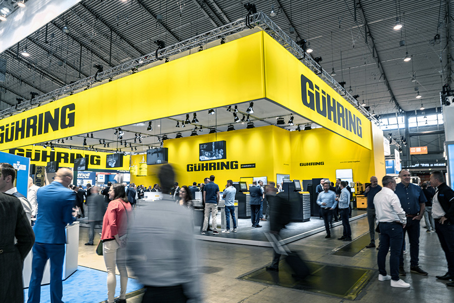Beschäftigte Messehalle mit einem markanten Gühring-Messestand in leuchtendem Gelb, der zahlreiche Besucher anzieht Beschäftigte Messehalle mit einem markanten Gühring-Messestand in leuchtendem Gelb, der zahlreiche Besucher anzieht