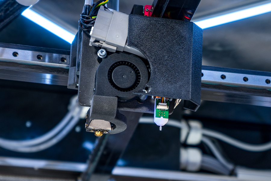 Industrieller 3D-Druck: Stabile Prozesse trotz abrasiver Filamente Abrasive Materialien bringen konventionelle Druckdüsen schnell an ihre Grenzen. Mit DIANOZ setzt Mach-3D auf eine verschleißstabile Lösung für reproduzierbare Druckprozesse – und integriert die Diamant-Düse künftig serienmäßig in seine Systeme. Mach-3D vereint mehrere additive Fertigungstechnologien unter einem Dach: Seit 2024 betreibt das Unternehmen vor den Toren Kiels FDM-, SLA- und SLS-Anlagen für die Prototypenfertigung und Serienproduktion. Ergänzend entwickelt und fertigt Mach-3D eigene industrielle 3D-Drucksysteme, mit denen Funktionsbauteile, Vorrichtungen, Gehäuse, Maschinenkomponenten und großformatige technische Bauteile gedruckt werden. Als Werkstoffe kommen dabei hauptsächlich technische Kunststoffe und glasfaserverstärkte Materialien zum Einsatz. Abrasive Materialien als Herausforderung Die Ansprüche sind hoch – vor allem an einen kleinen, aber zentralen Teil des 3D-Druckers: Die Druckerdüse. „Für uns stehen vor allem Prozesssicherheit, Verschleißfestigkeit und eine gleichbleibend hohe Druckqualität im Vordergrund“, erklärt Erik Dieckmann, Geschäftsführer von Mach-3D. „Die Düse muss auch bei abrasiven Werkstoffen über lange Zeit ein konstantes Austragsverhalten gewährleisten. Darüber hinaus erwarten wir eine hohe Maßhaltigkeit der Bauteile, saubere Oberflächen und die Möglichkeit, auch größere Materialdurchsätze für industrielle Anwendungen zuverlässig zu realisieren.“ Und genau da liegt oft der Knackpunkt, wie Dieckmann aus Erfahrung weiß: „Die größte Herausforderung liegt in der hohen Abrasivität glasfaserverstärkter Materialien. Herkömmliche Düsen verschleißen hier relativ schnell.“ In der Praxis führte das bei Mach-3D häufig zu instabilen Prozessen. Der Düsendurchmesser änderte sich mit der Laufzeit und damit auch das Extrusionsverhalten der Düse. Die Folge: Qualitätsschwankungen, zusätzlicher Prüfaufwand und ungeplante Stillstände. Besonders bei großformatigen Bauteilen mit langen Druckzeiten von Stunden oder sogar Tagen sowie bei Projekten mit hohen Anteilen glasfaserverstärkter Materialien wurden die Grenzen herkömmlicher Düsen deutlich. Klassische Lösungen stoßen an Grenzen Mach-3D versuchte, mit Stahldüsen und Rubindüsen das Verschleißproblem beim Druck abrasiver Materialien in den Griff zu bekommen. Beide Ansätze lieferten jedoch keine stabile Lösung für den industriellen Dauereinsatz. Bei den Stahldüsen trat bei längerem Einsatz Verschleiß auf, der die Druckqualität beeinflusste. Rubindüsen boten zwar eine hohe Verschleißfestigkeit, waren jedoch empfindlicher gegenüber mechanischen Belastungen. „Für den laufenden Betrieb bedeutete dies zusätzlichen Wartungsaufwand und teilweise ungeplante Stillstände“, so Deickmann. Entscheidend war daher die Suche nach einer Düse, die nicht nur länger hält, sondern dabei auch konstante Ergebnisse liefert. DIANOZ bringt Prozessstabilität zurück Mit der Diamantdüse DIANOZ von Gühring gelang der Wechsel zu einer stabilen Lösung. Was diese Düse so besonders macht: Die DIANOZ verfügt über eine Spitze aus polykristallinem Diamant (PKD) mit deutlich höherer Härte als Messing oder gehärteter Stahl, wodurch Geometrie und Austrittsdurchmesser auch bei abrasiven Filamenten stabil bleiben. Der Filamentkanal ist reibungsoptimiert und im konischen Engbereich diamantverstärkt ausgeführt, um Verschleiß und Strömungsstörungen zu minimieren. Aufgrund der hohen Wärmeleitfähigkeit des Diamanten entspricht die tatsächliche Düsentemperatur weitgehend der Solltemperatur, was zu reproduzierbaren thermischen Bedingungen im Schmelzbereich führt. Für Mach-3D zeigt sich der Nutzen im Betrieb: „Der größte Vorteil liegt für uns in der Kombination aus hoher Verschleißfestigkeit, Prozesssicherheit und gleichbleibender Druckqualität“, sagt Erik Dieckmann. DIANOZ liefere ihm endlich die Zuverlässigkeit, nach der er gesucht hatte. „Bereits nach den ersten Druckversuchen war erkennbar, dass die Düse für den industriellen Einsatz entwickelt wurde“, berichtet Dieckmann. Besonders überzeugt habe ihn die Kombination aus hochwertiger Verarbeitung, technischer Auslegung für abrasive Materialien und der Erfahrung von Gühring als Zerspanungsspezialist. Konstante Qualität über lange Laufzeiten Die Wirkung wird vor allem im Langzeiteinsatz sichtbar. Während sich bei konventionellen Düsen das Druckbild verändert, bleibt DIANOZ stabil. „Auch nach der Verarbeitung abrasiver Materialien konnten wir keine relevanten Veränderungen hinsichtlich Druckbild, Materialfluss oder Bauteilqualität feststellen“, bestätigt Dieckmann. Diese Stabilität verbessert die Reproduzierbarkeit. Maßhaltigkeit und Oberflächenqualität bleiben konstant, unabhängig von der Laufzeit. Gleichzeitig sinkt der Wartungsaufwand. „Wir konnten den Aufwand für Kontrolle, Wartung und Düsenwechsel spürbar reduzieren“, erklärt Dieckmann. Die Maschinen laufen länger ohne Eingriff, die Produktion lässt sich besser planen. Partnerschaft statt Lieferantenbeziehung Mittlerweile setzt Mach-3D die Diamantdüse als Standard in der eigenen Produktion ein. „Aus einer klassischen Lieferantenbeziehung hat sich eine enge technologische Partnerschaft entwickelt“, beschreibt Dieckmann. Dabei schätzt der Geschäftsführer an Gühring vor allem, dass der Werkzeughersteller zwei entscheidende Faktoren vereint: hervorragende Produktqualität und jahrzehntelange Erfahrung im Bereich hochverschleißfester Präzisionswerkzeuge. „Während viele Anbieter ausschließlich aus dem 3D-Druck-Umfeld kommen, bringt Gühring umfangreiches Know-how aus der Zerspanung und Werkstofftechnik mit“, stellt Dieckmann fest. „Diese Kompetenz spiegelt sich unmittelbar in der Leistungsfähigkeit der DIANOZ-Düsen wider.“ Die Zusammenarbeit mündet nun in eine OEM-Partnerschaft. Mach-3D wird DIANOZ serienmäßig in seinen neuen Großformatdruckern einsetzen. „Für unsere neue Maschine war klar, dass wir unseren Kunden auch bei der Druckerdüse von Beginn an eine bewährte Lösung anbieten möchten“, begründet Dieckmann diese Entscheidung. DIANOZ erfüllt die hohen Anforderungen der Industrie, und in Kombination mit den Großformatdruckern von Mach-3D entsteht ein abgestimmtes System für die Anwender. Industrieller 3D-Druck mit DIANOZ-Diamantdüse von Gühring zur Verarbeitung abrasiver Filamente und glasfaserverstärkter Kunststoffe mit hoher Prozesssicherheit.